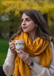 a beautiful young girl sits on the grass in an autumn park, holds a cup in her hands and looks away. Reflections, enjoyment